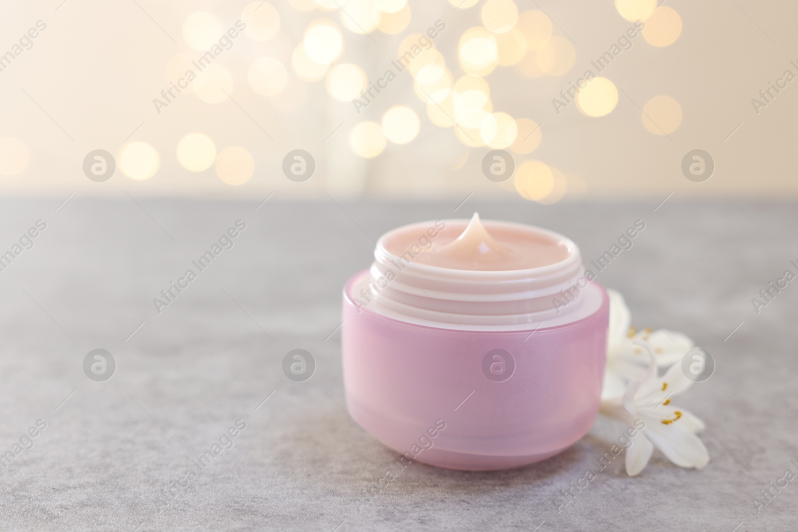 Photo of Cream in jar and beautiful jasmine flowers on grey table against light background with blurred lights, closeup