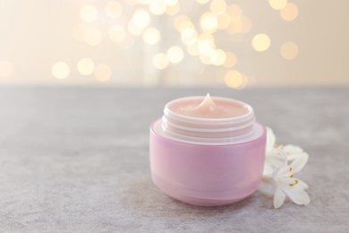 Photo of Cream in jar and beautiful jasmine flowers on grey table against light background with blurred lights, closeup