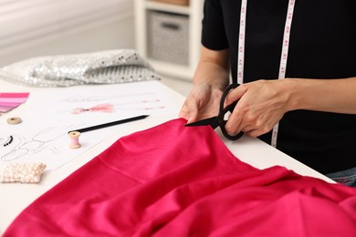 Photo of Fashion designer cutting pink fabric at table in workshop, closeup