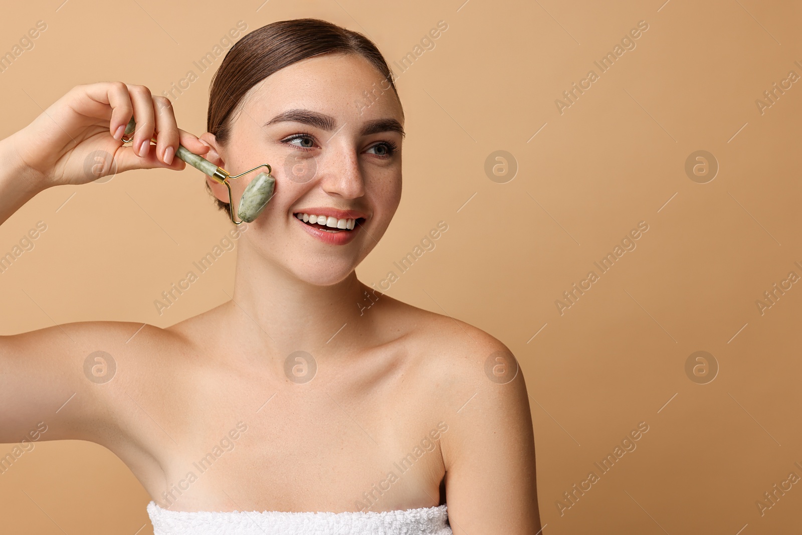 Photo of Beautiful young woman doing facial massage with roller on beige background, space for text