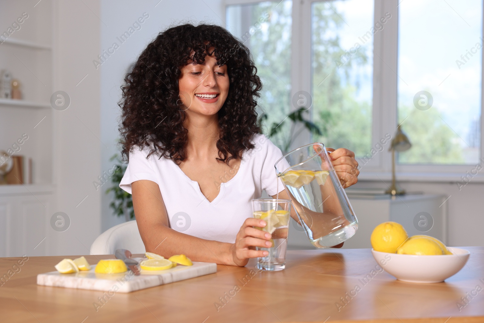 Photo of Woman pouring lemon water from jug into glass at table indoors