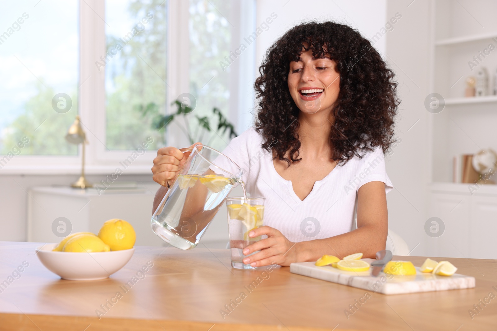 Photo of Woman pouring lemon water from jug into glass at table indoors