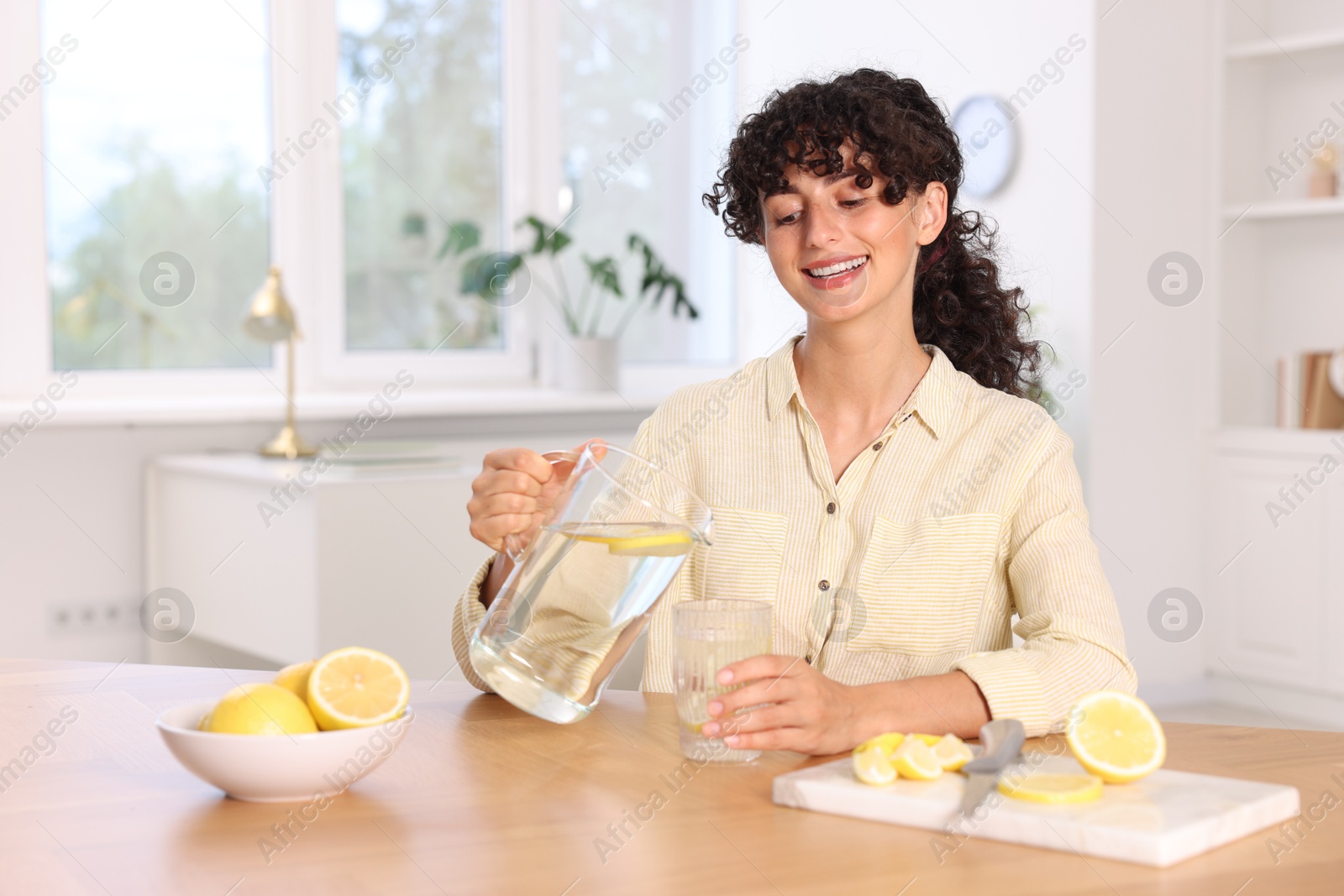 Photo of Woman pouring lemon water from jug into glass at table indoors