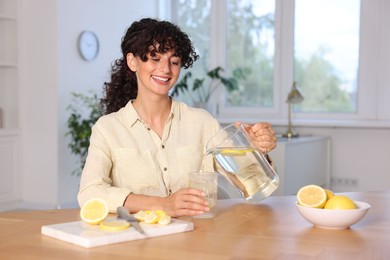 Photo of Woman pouring lemon water from jug into glass at table indoors