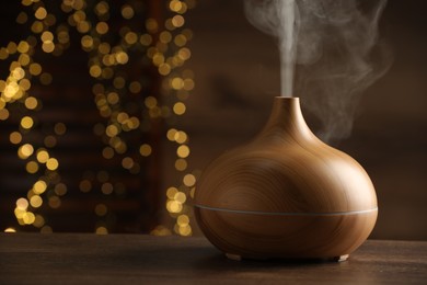 Photo of Aroma oil diffuser on wooden table against blurred lights, space for text