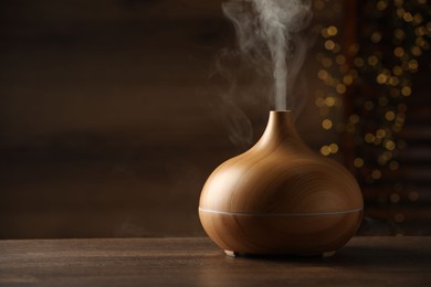 Photo of Aroma oil diffuser on wooden table against blurred lights, space for text