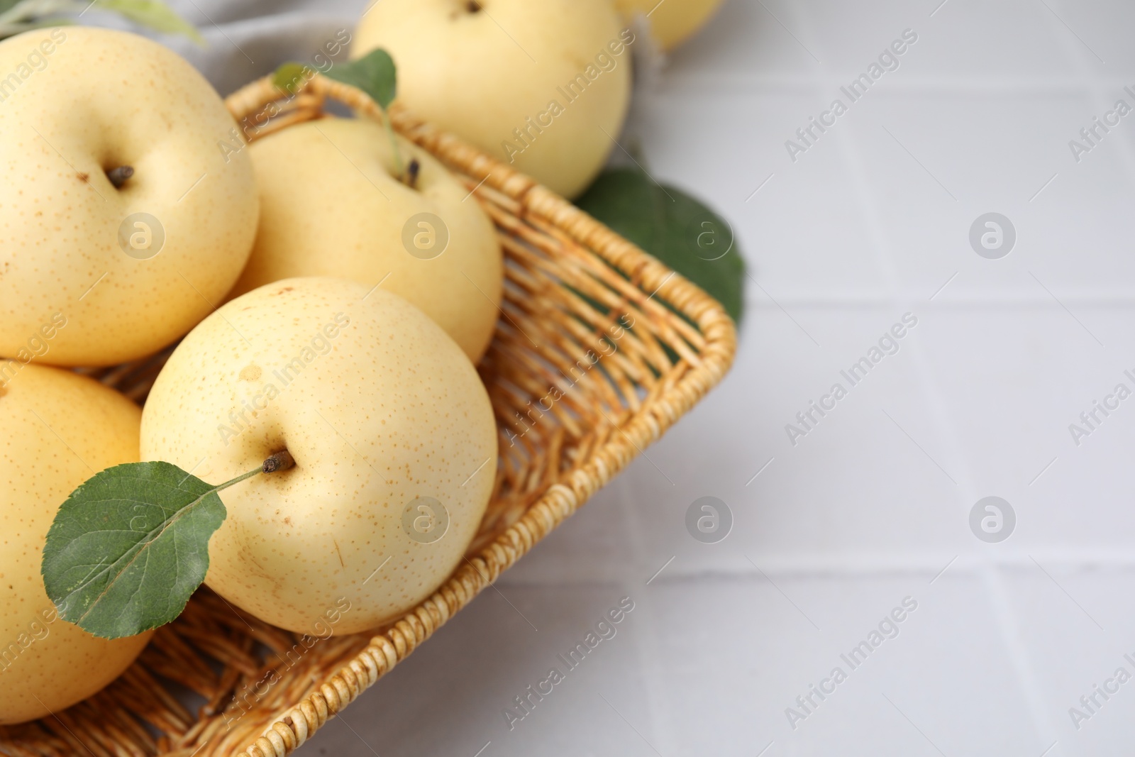 Photo of Delicious fresh apple pears in wicker basket and green leaves on white tiled table, closeup. Space for text