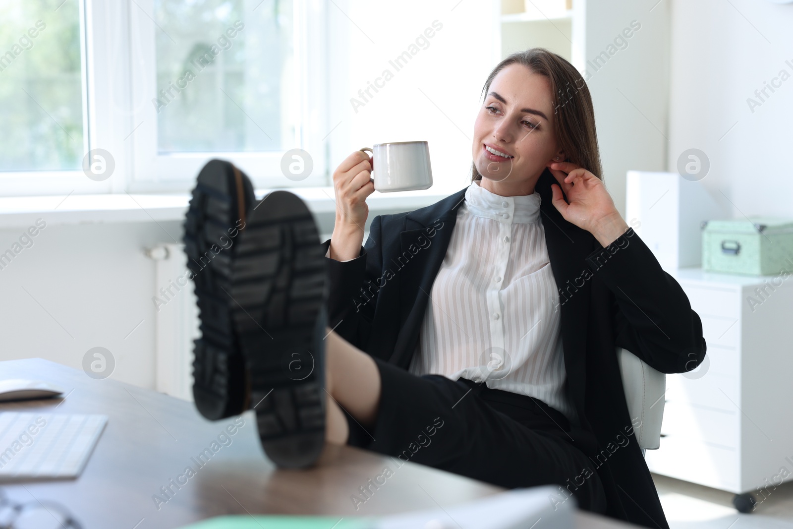 Photo of Smiling businesswoman drinking coffee holding legs on table in office. Break time