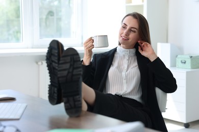 Photo of Smiling businesswoman drinking coffee holding legs on table in office. Break time