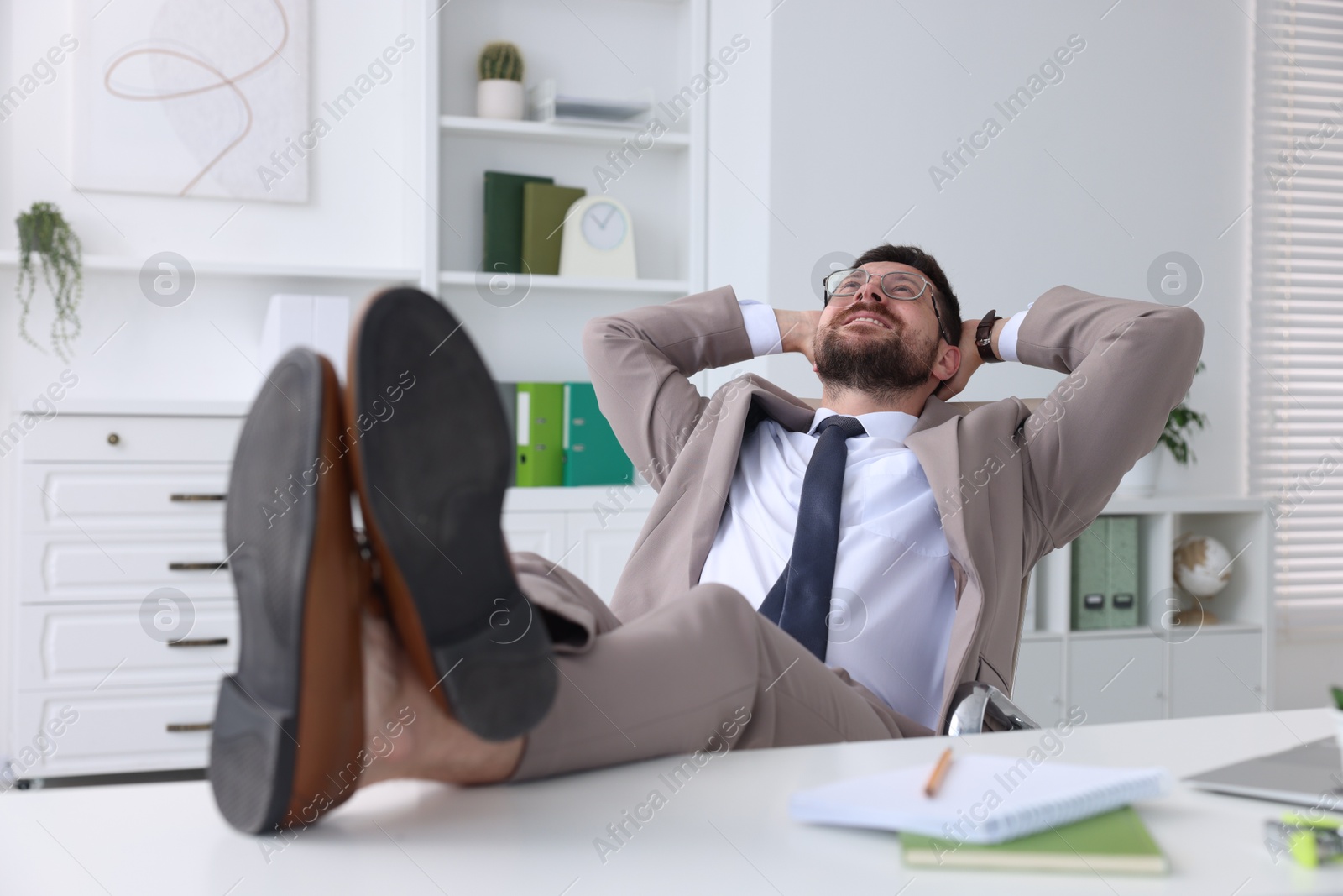 Photo of Smiling businessman with hands behind his head holding legs on table in office. Break time