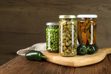 Photo of Different pickled products in jars and fresh jalapeno peppers on wooden table