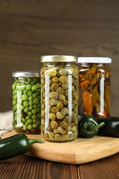 Photo of Different pickled products in jars and fresh jalapeno peppers on wooden table