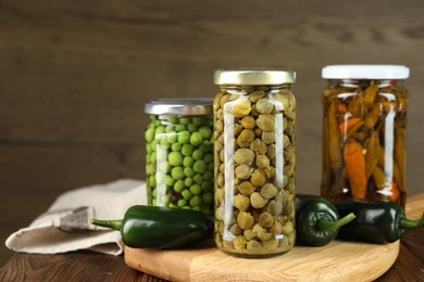 Photo of Different pickled products in jars and fresh jalapeno peppers on wooden table