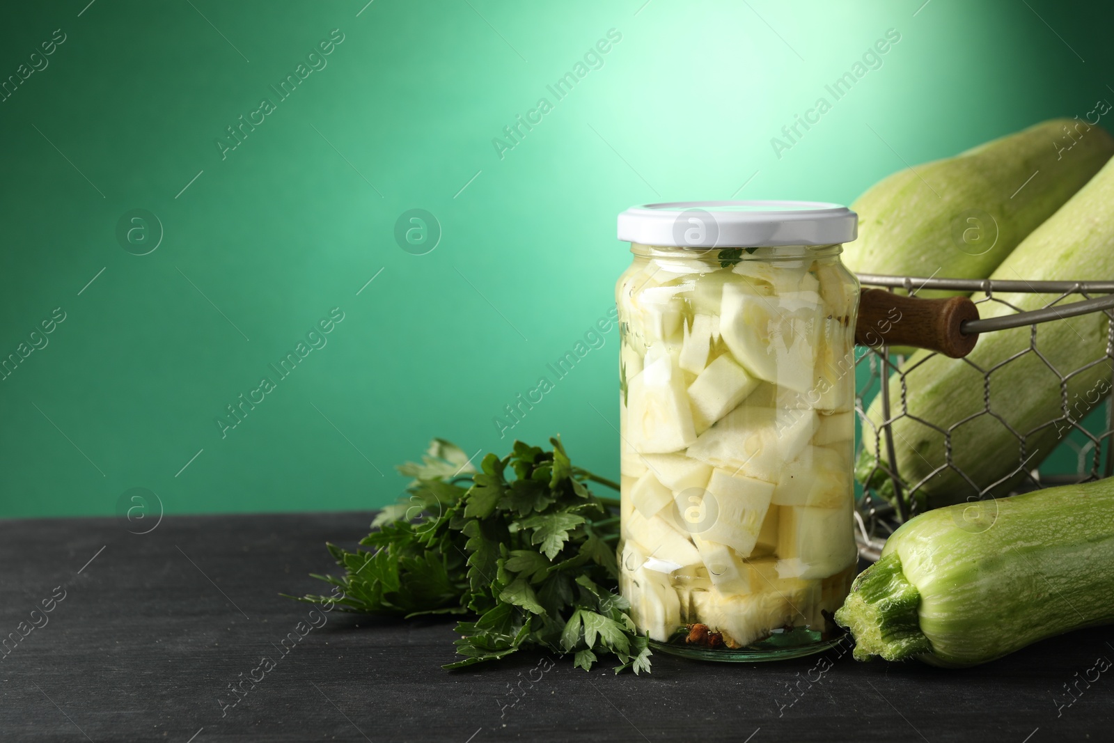 Photo of Tasty pickled zucchini in jar, fresh vegetables and parsley on black wooden table, space for text