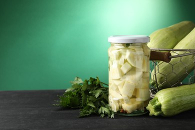 Photo of Tasty pickled zucchini in jar, fresh vegetables and parsley on black wooden table, space for text