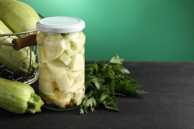 Photo of Tasty pickled zucchini in jar, fresh vegetables and parsley on black wooden table, space for text