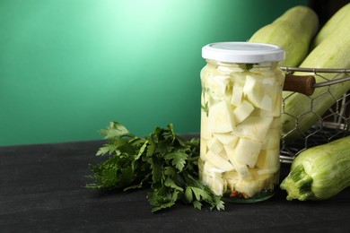 Photo of Tasty pickled zucchini in jar, fresh vegetables and parsley on black wooden table