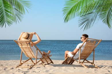 Couple relaxing on deck chairs under palm leaves on tropical beach