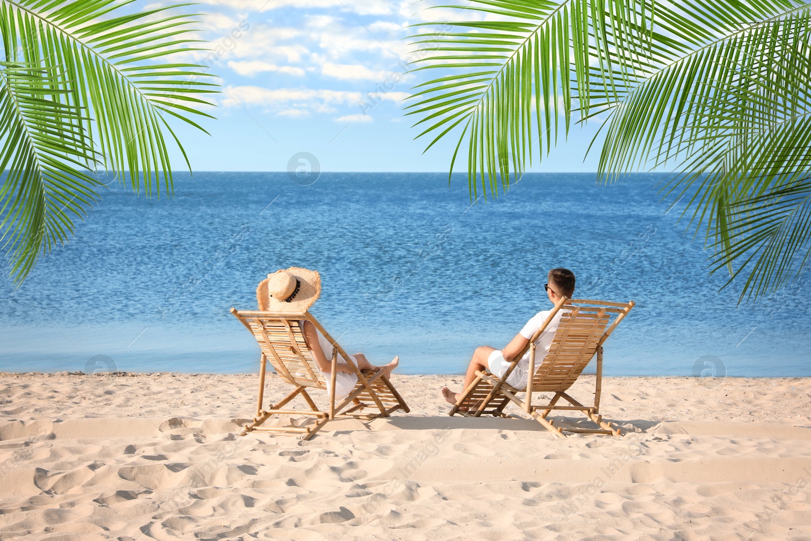 Image of Couple relaxing on deck chairs under palm leaves on tropical beach