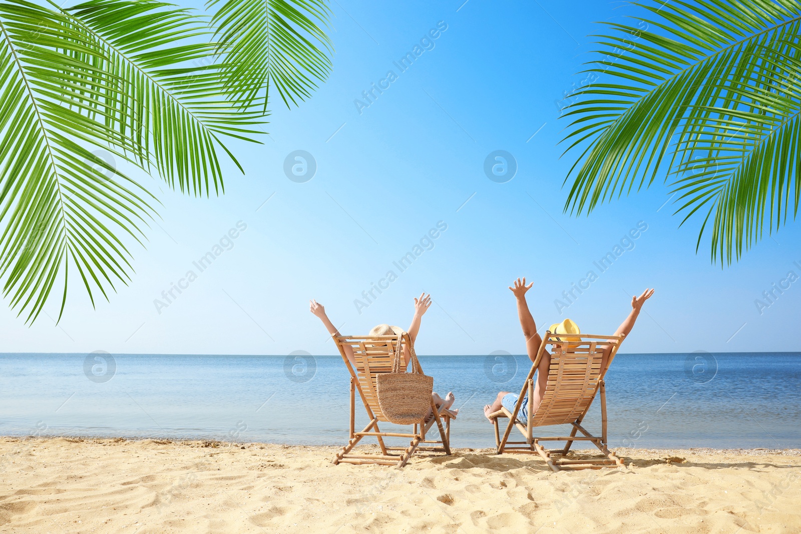 Image of Couple relaxing on deck chairs under palm leaves on tropical beach