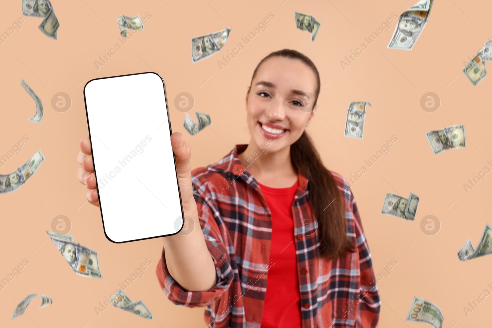 Image of Happy woman showing mobile phone with empty screen under money rain on dark beige background