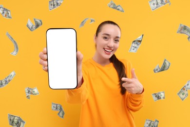 Image of Happy woman pointing at mobile phone with empty screen under money rain on orange background