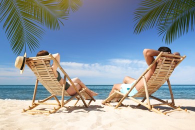 Image of Couple relaxing on deck chairs under palm leaves on tropical beach