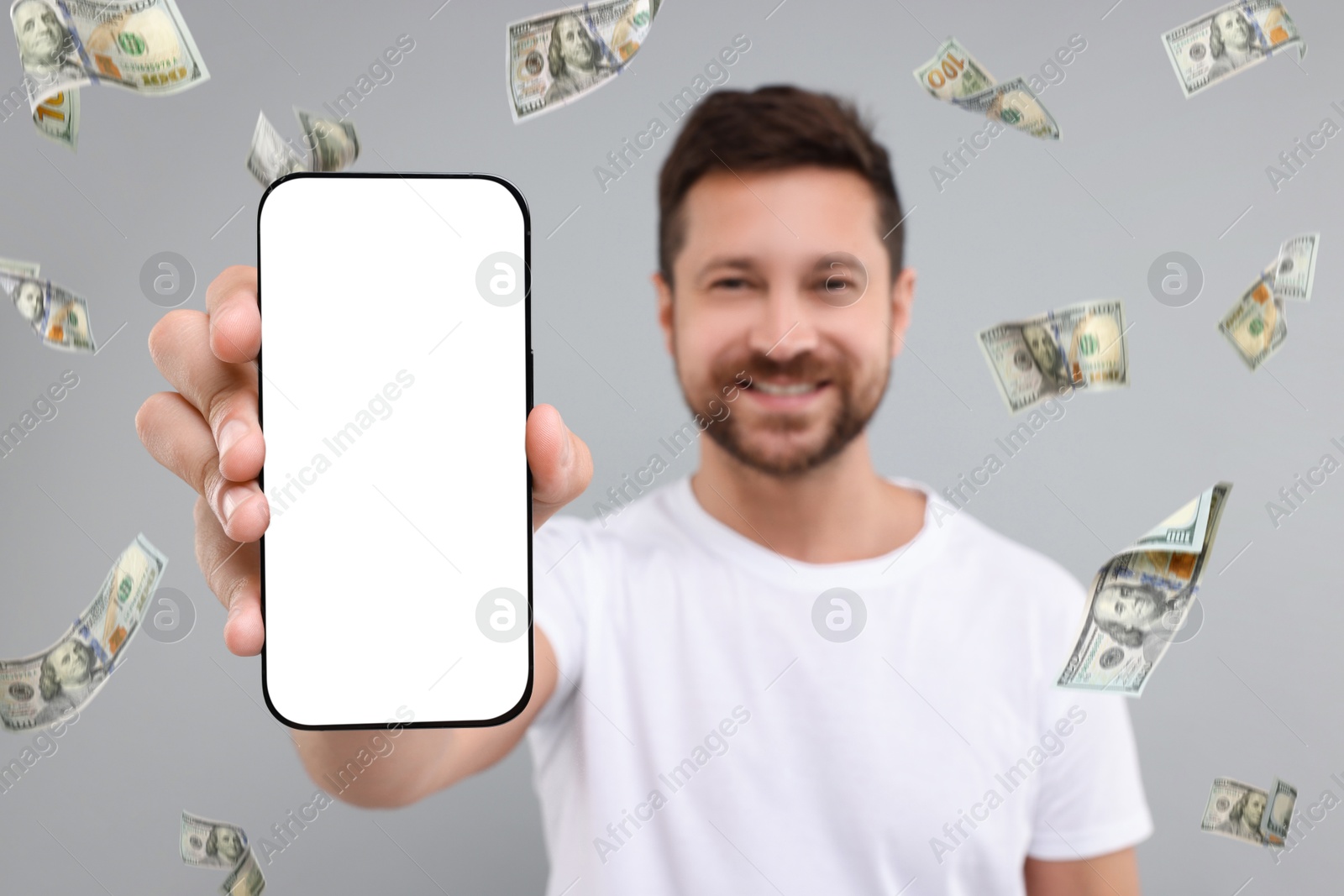 Image of Happy man showing mobile phone with empty screen under money rain on grey background, selective focus