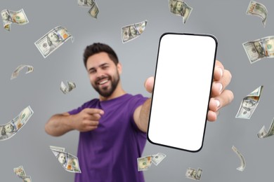 Happy man pointing at mobile phone with empty screen under money rain on grey background, selective focus