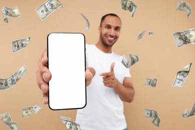 Happy man pointing at mobile phone with empty screen under money rain on dark beige background, selective focus