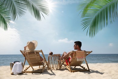 Couple relaxing on deck chairs under palm leaves on tropical beach