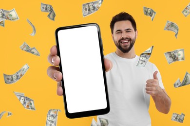 Happy man showing mobile phone with empty screen under money rain on orange background