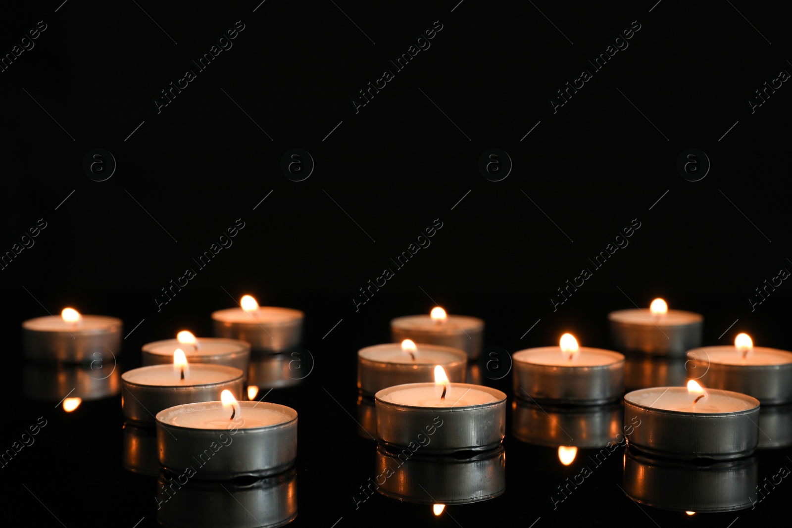 Photo of Many burning tealight candles on mirror surface against black background