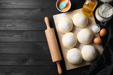 Raw dough balls, yolk, flour, eggs and rolling pin on black wooden table, top view. Space for text