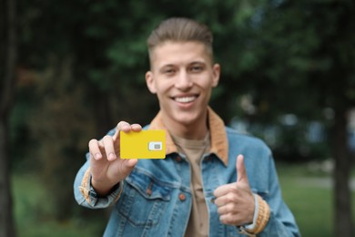 Photo of Happy man with SIM card showing thumbs up outdoors, selective focus