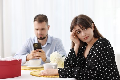 Photo of Embarrassing date. Bored woman sitting at table with man using smartphone indoors, selective focus