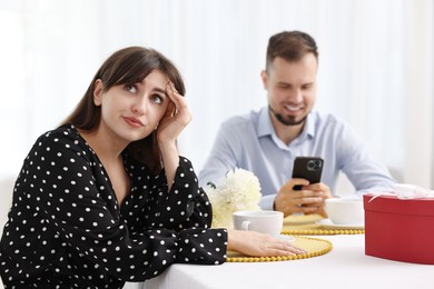 Photo of Embarrassing date. Bored woman sitting at table with man using smartphone indoors, selective focus