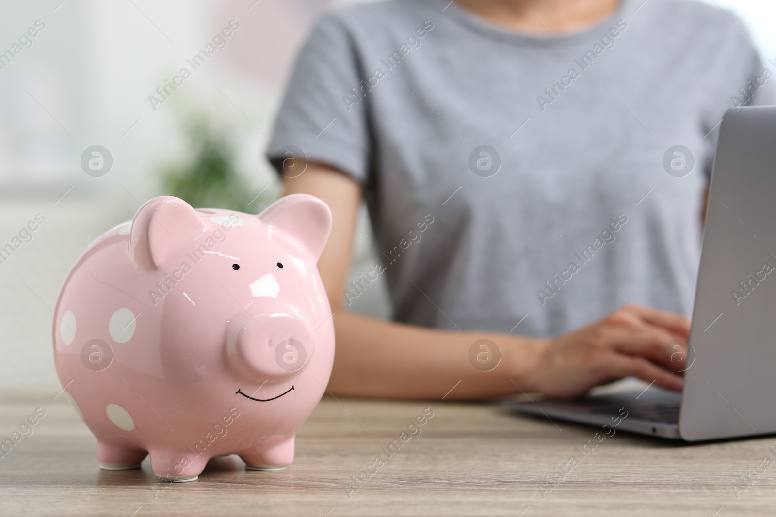 Photo of Woman using laptop at wooden table, focus on piggy bank indoors