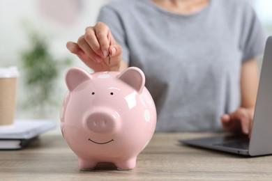 Photo of Woman putting coin into piggy bank while using laptop at wooden table, closeup