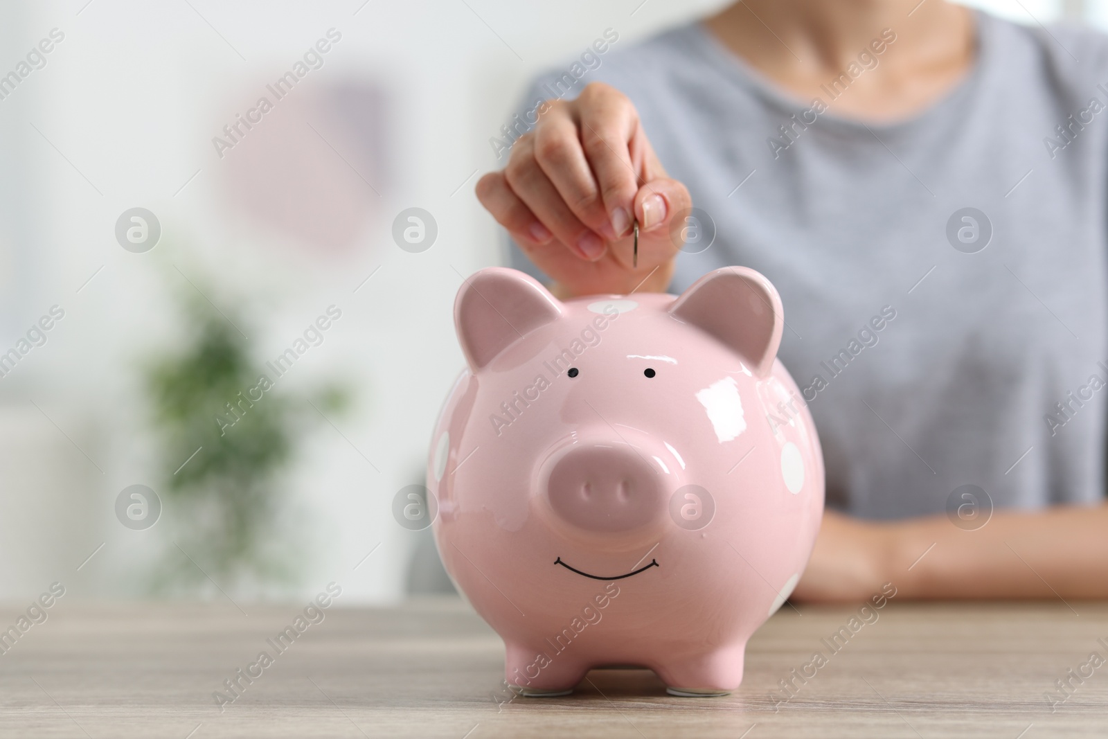 Photo of Woman putting coin into piggy bank at wooden table, closeup