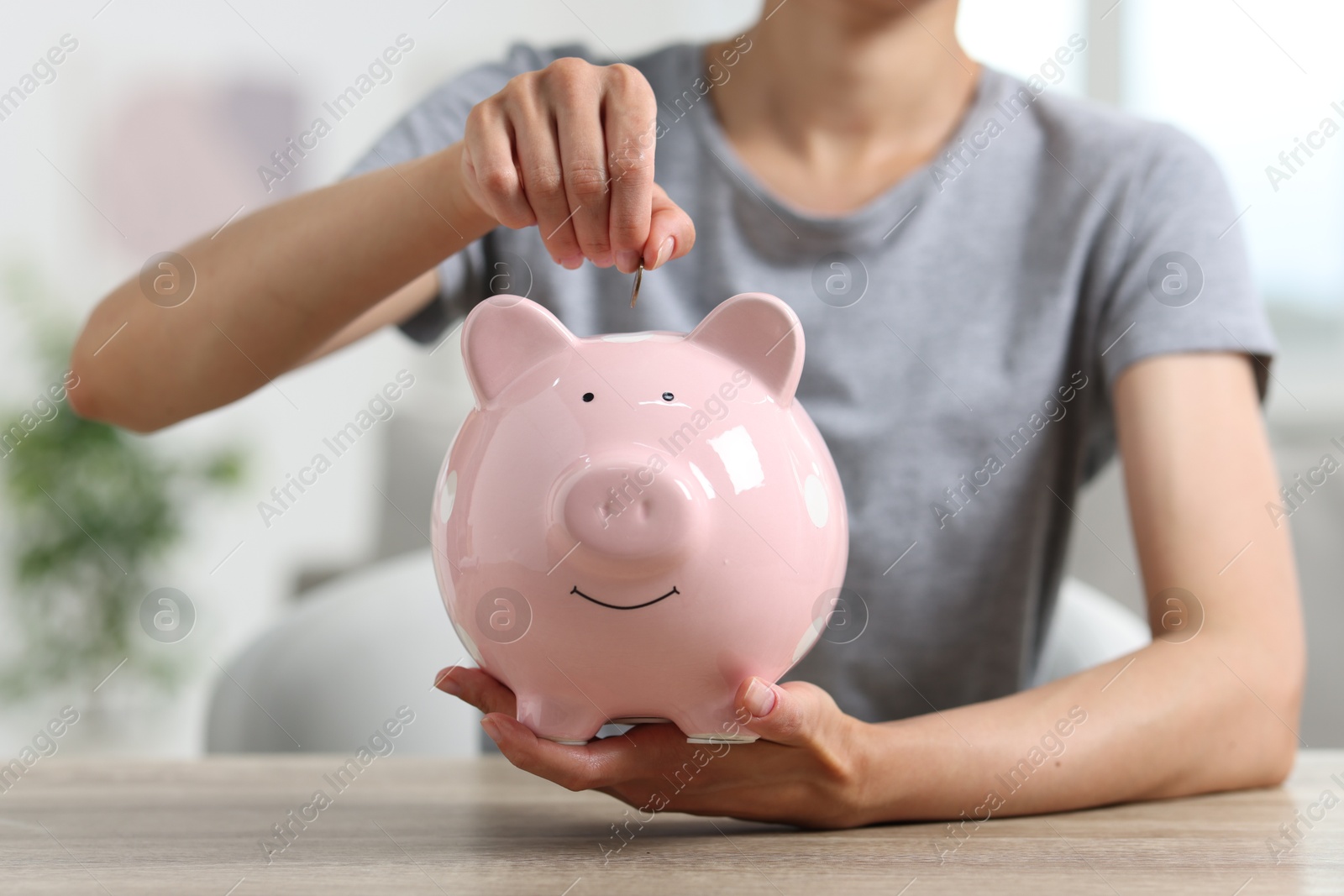 Photo of Woman putting coin into piggy bank at wooden table, closeup