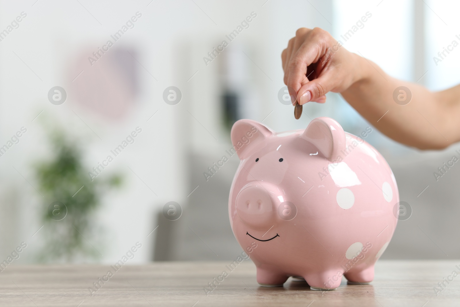 Photo of Woman putting coin into piggy bank at wooden table, closeup. Space for text