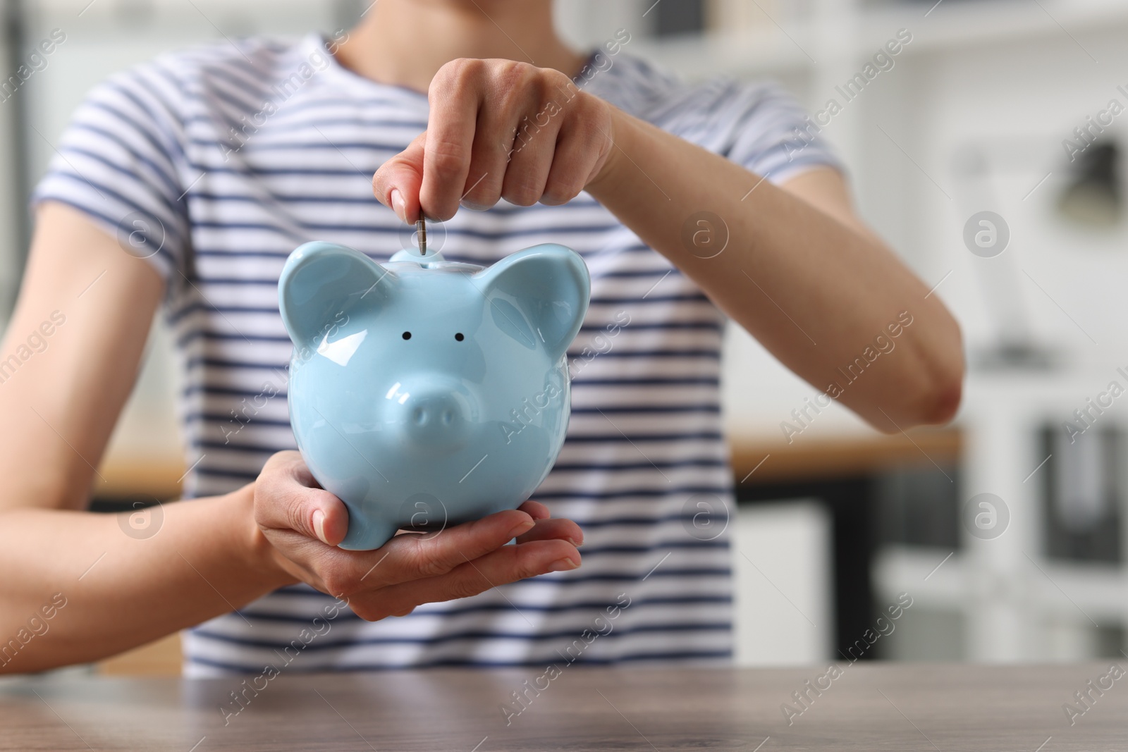 Photo of Woman putting coin into piggy bank at wooden table, closeup