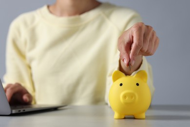 Photo of Woman putting coin into piggy bank while using laptop at table, closeup