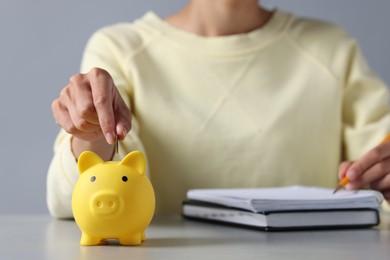 Photo of Woman putting coin into piggy bank while taking notes at table, closeup