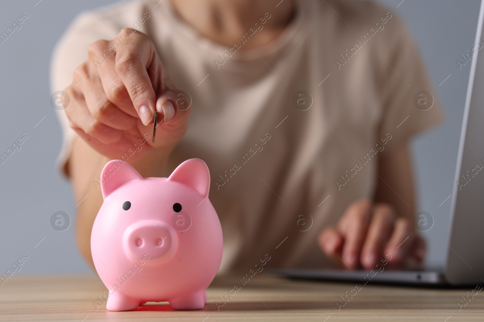 Photo of Woman putting coin into piggy bank while using laptop at wooden table, closeup