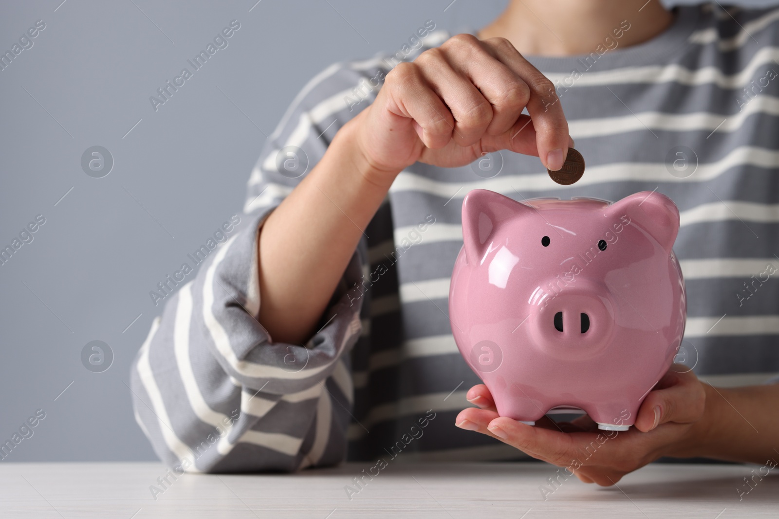 Photo of Woman putting coin into piggy bank at white wooden table, closeup