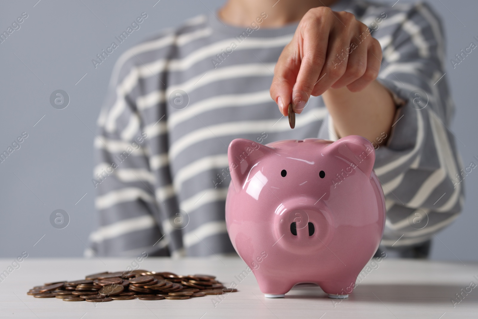 Photo of Woman putting coin into piggy bank at white wooden table, closeup