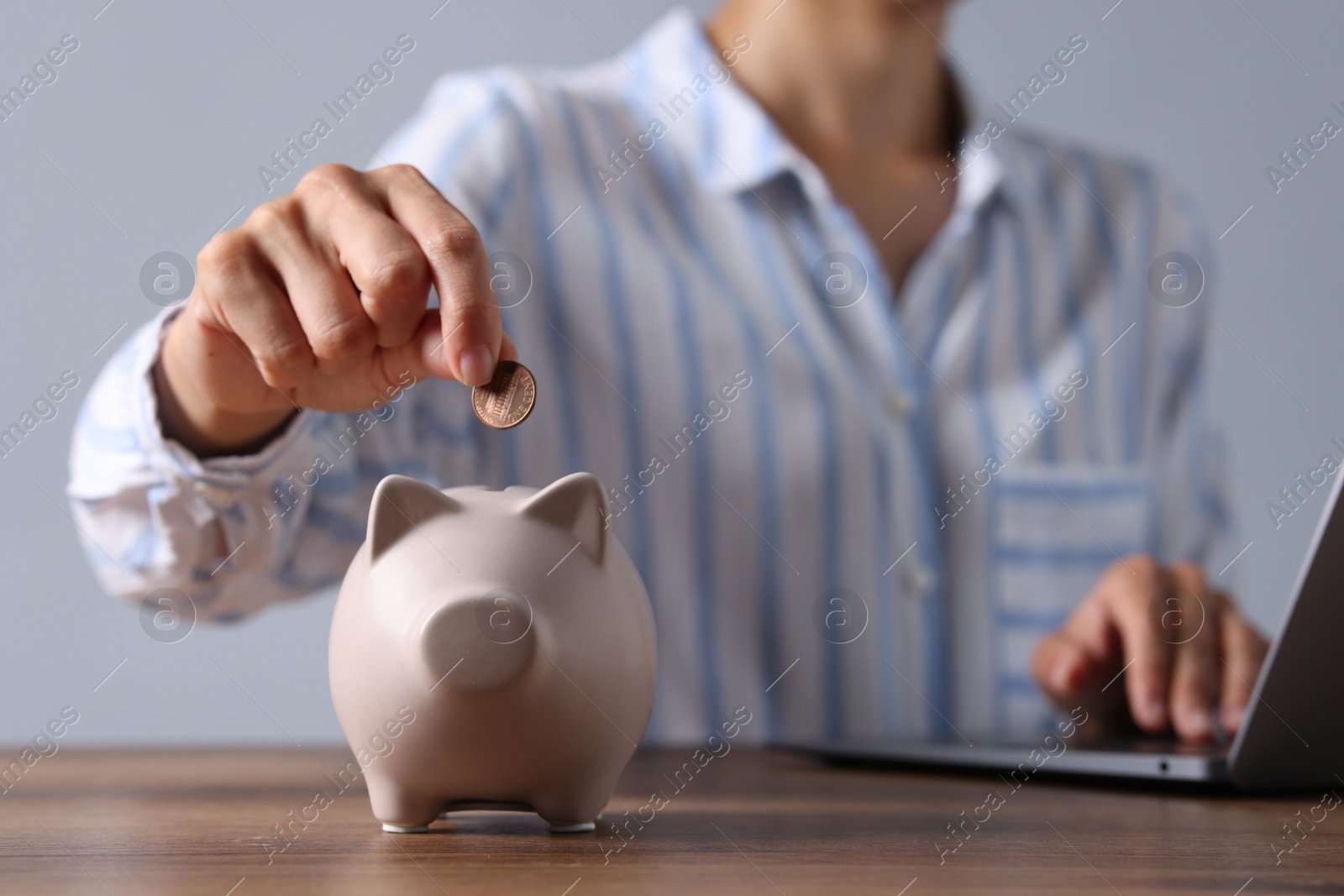 Photo of Woman putting coin into piggy bank while using laptop at wooden table, closeup