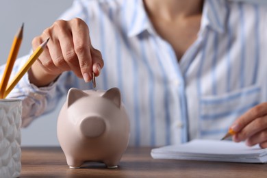 Woman putting coin into piggy bank while taking notes at wooden table, closeup